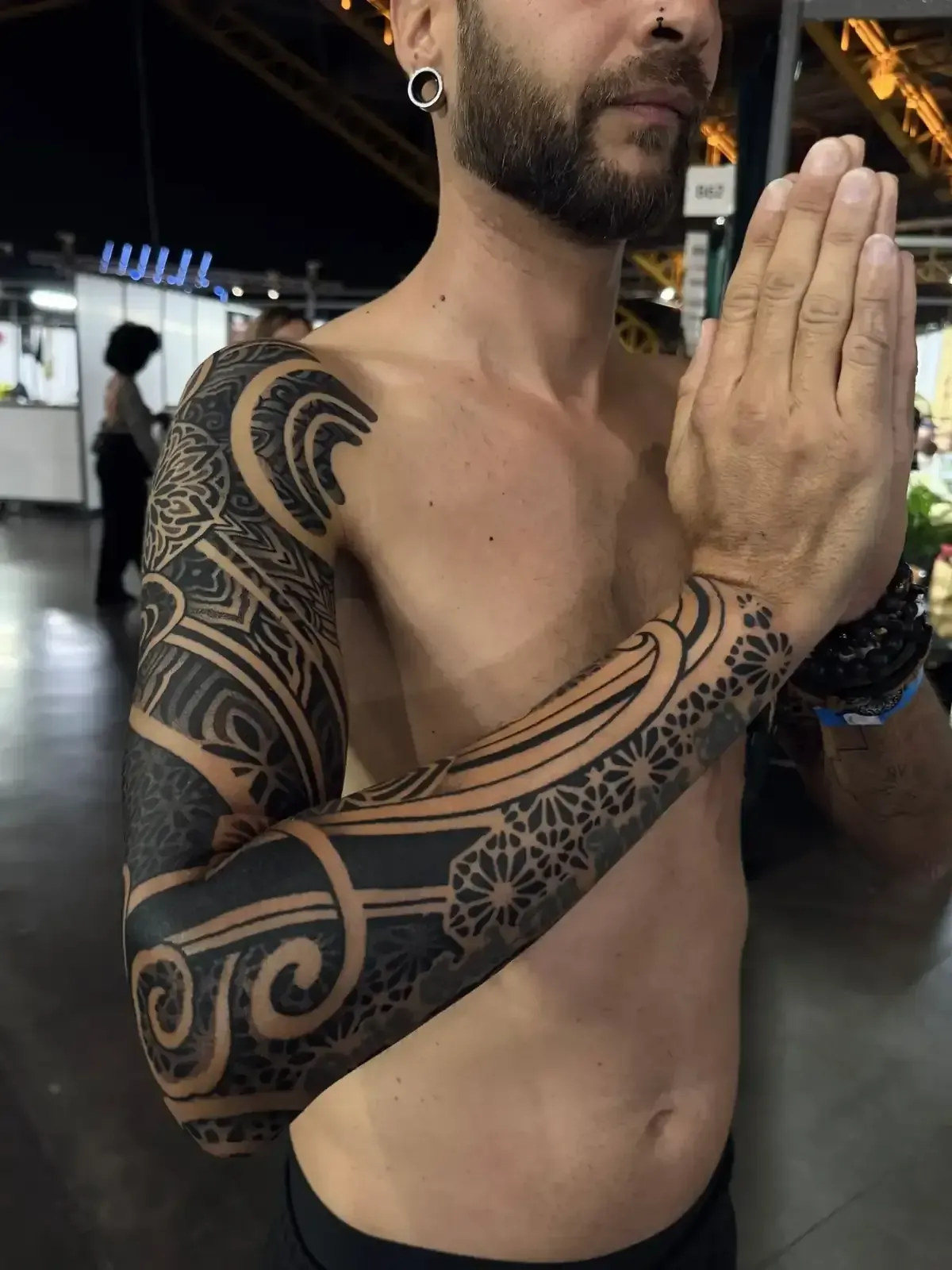 Hombre con tatuajes en las mangas, con las manos juntas en postura de oración. Entorno oscuro, piel clara.
