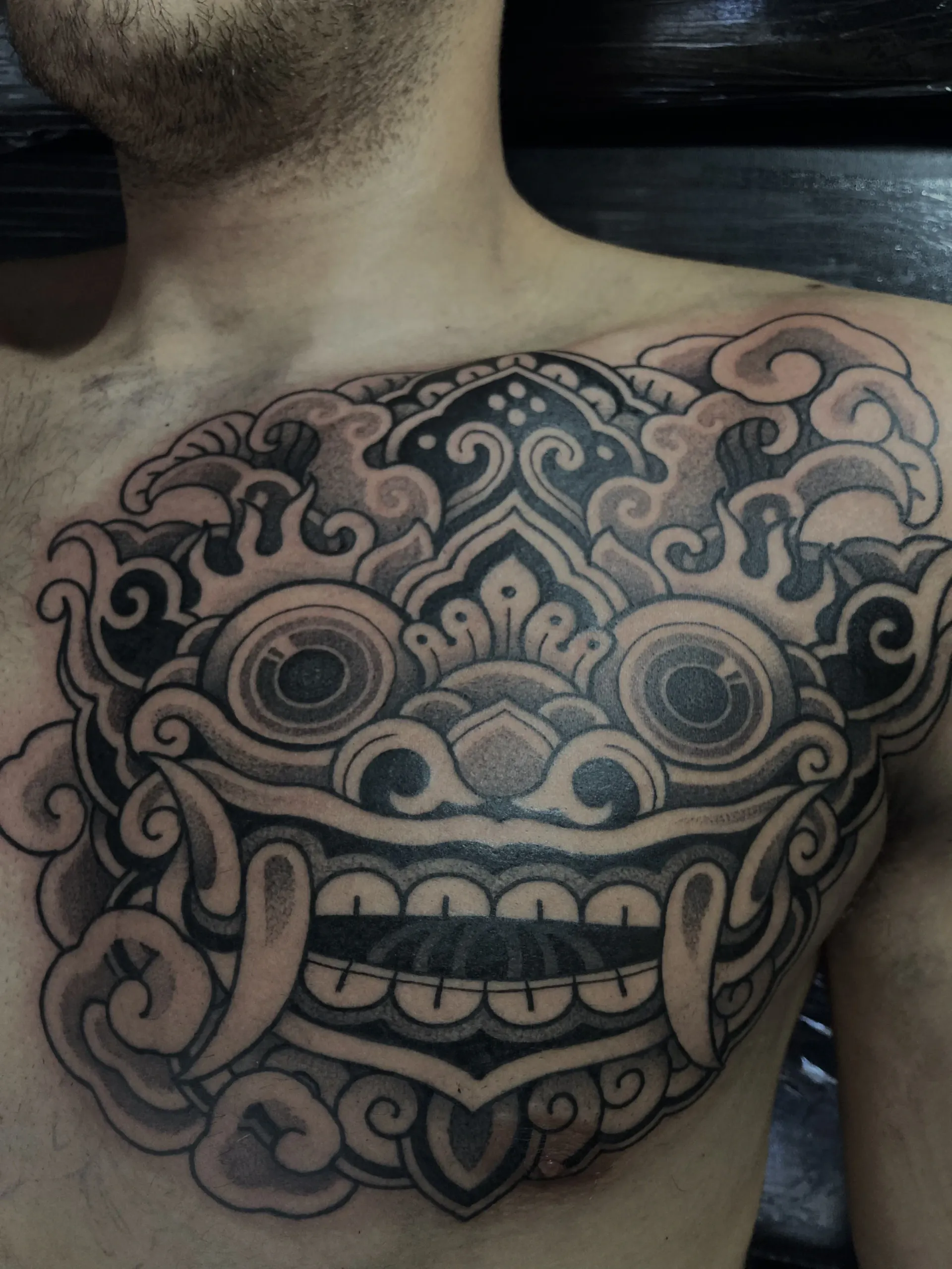 Tatuaje en el pecho, en color negro y gris, de una máscara Barong balinesa estilizada.