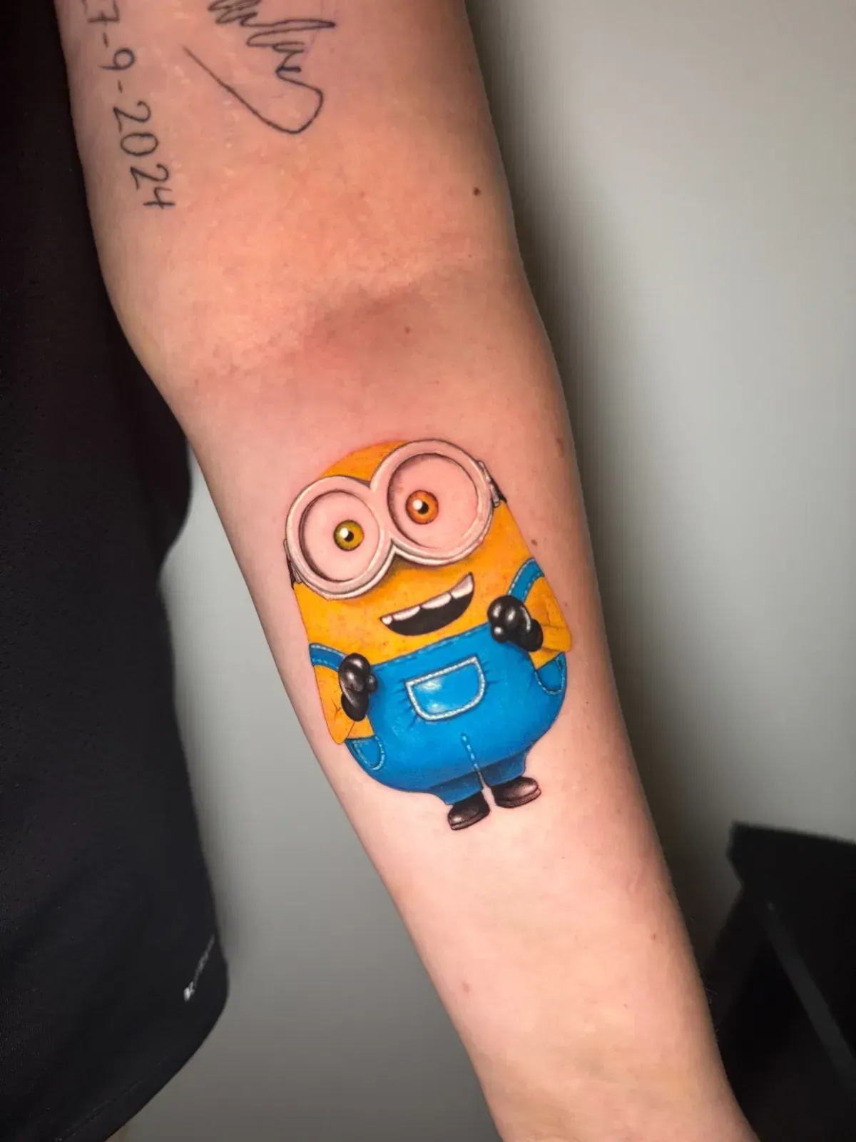 Tatuaje de Minion en el antebrazo; cuerpo amarillo, mono azul, ojos grandes, guantes negros.