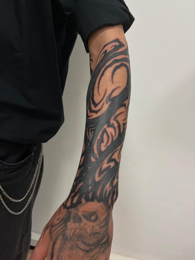 Brazo con tatuaje tribal negro en la manga y tatuaje parcial en el antebrazo. La persona lleva camisa negra.