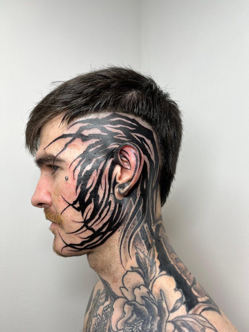 Hombre con extensos tatuajes negros en la cara y el cuello, cabello corto, perfil lateral.