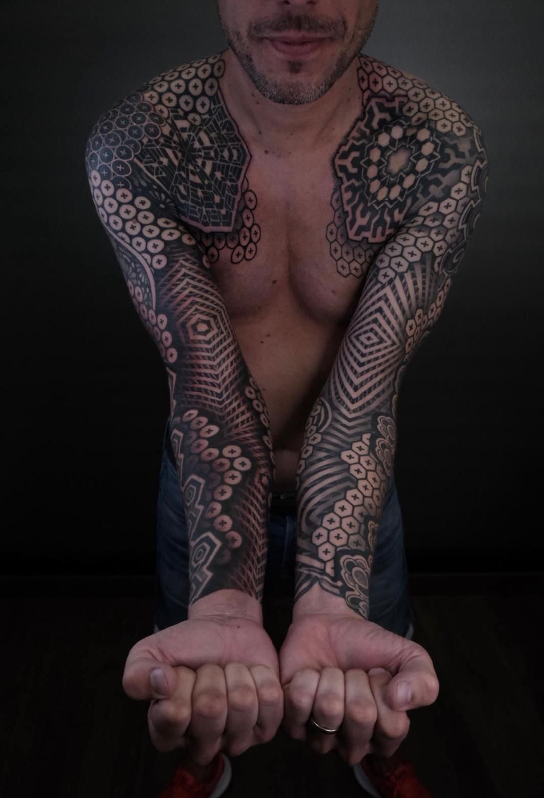 Hombre con intrincados tatuajes en el brazo en blanco y negro, con las manos abiertas; fondo oscuro.