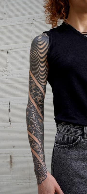 Primer plano de un brazo tatuado con un tatuaje geométrico negro en la manga, junto a una camisa negra y pantalones vaqueros grises.