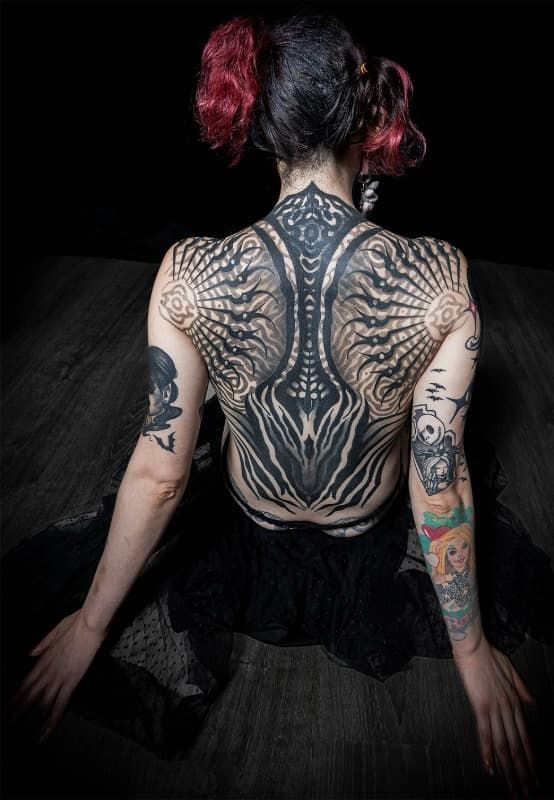 Vista posterior de una persona tatuada con cabello oscuro y trenzas teñidas de rojo, sentada contra un fondo oscuro.