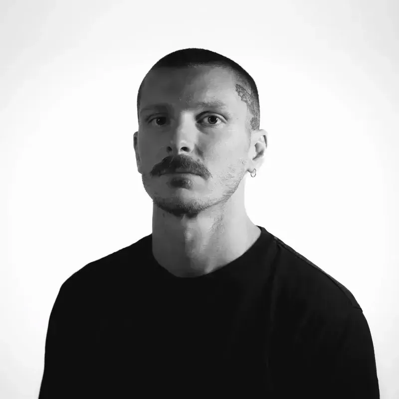 Hombre con bigote, cabeza rapada y camiseta negra, mirando al espectador.