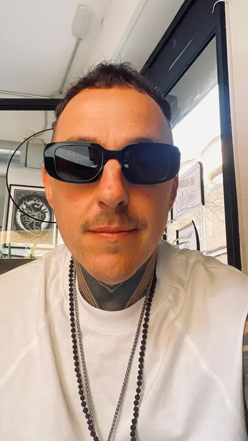 Hombre con gafas de sol y camisa blanca, con un tatuaje en el cuello y collares de cuentas. Interior.