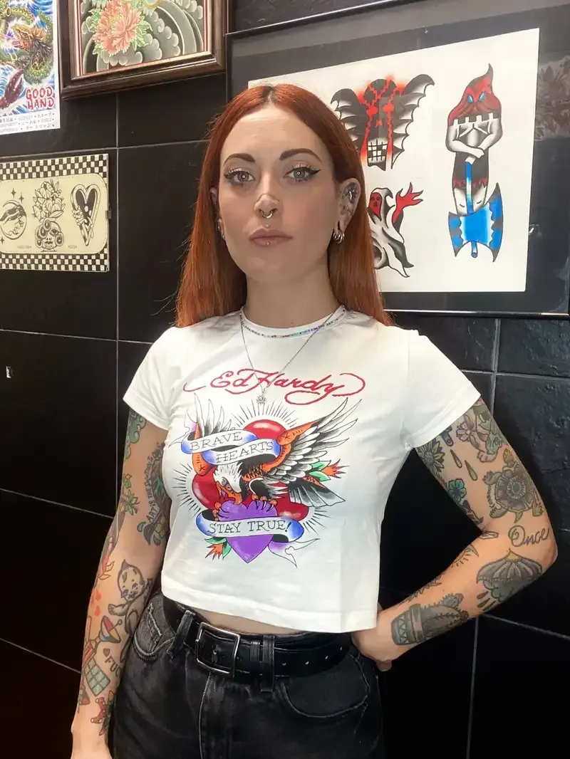 Mujer con cabello rojo y tatuajes, vistiendo una camiseta blanca, parada en un salón de tatuajes, con arte en las paredes.