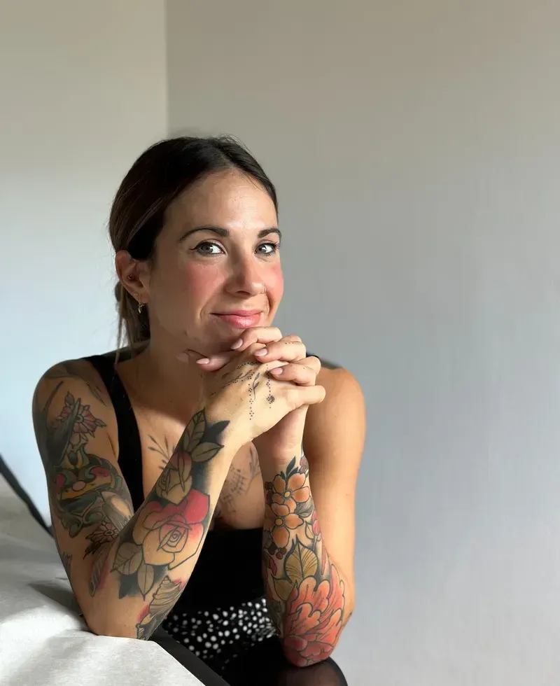 Una mujer con tatuajes en el brazo apoya la barbilla sobre sus manos entrelazadas, sonriendo.