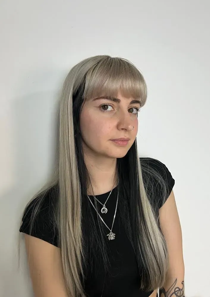 Mujer con cabello largo rubio y negro, flequillo, camisa negra y collares plateados.