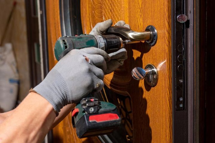 Persona con guantes que utiliza un taladro eléctrico para instalar una manija en una puerta de madera.