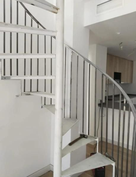 Escalera de caracol con escalones blancos y barandillas metálicas, que conduce a una zona de cocina.