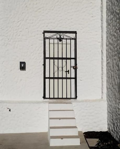 Puerta de seguridad de metal negro con rejas sobre una puerta blanca, juego de escalones blancos, pared texturizada blanca.