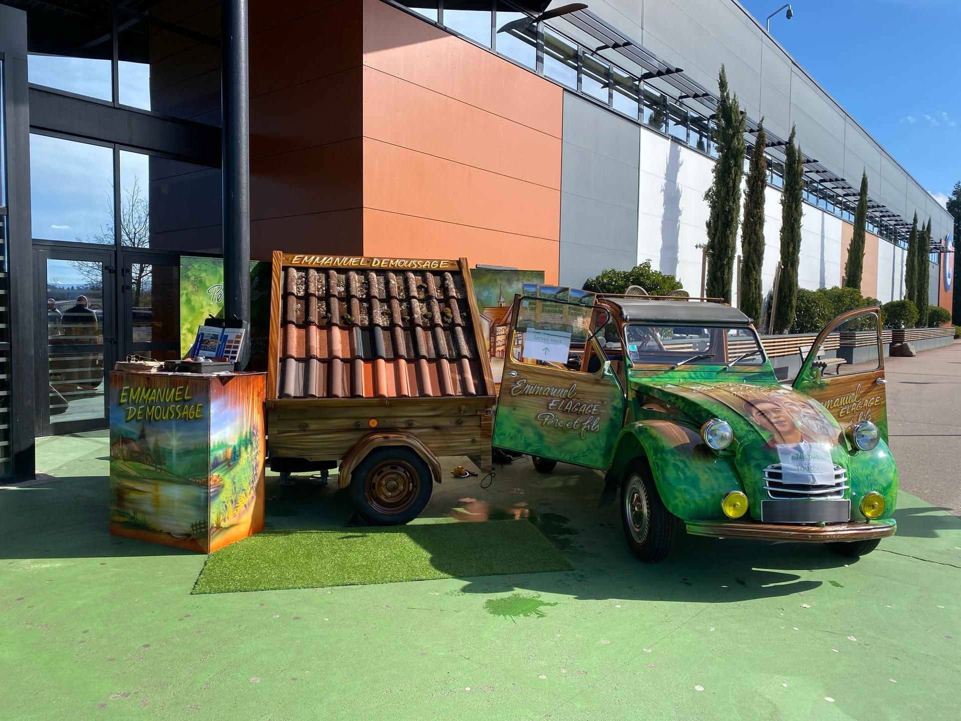 Voiture verte avec remorque, à côté d'un bâtiment aux panneaux orange. Installation d'exposition lors d'un événement.