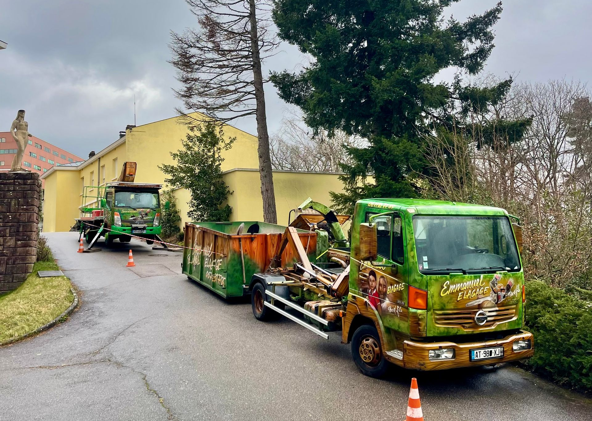 Deux camions verts chargés de débris se trouvent sur une route devant un bâtiment jaune ; un arbre est en train d'être abattu.