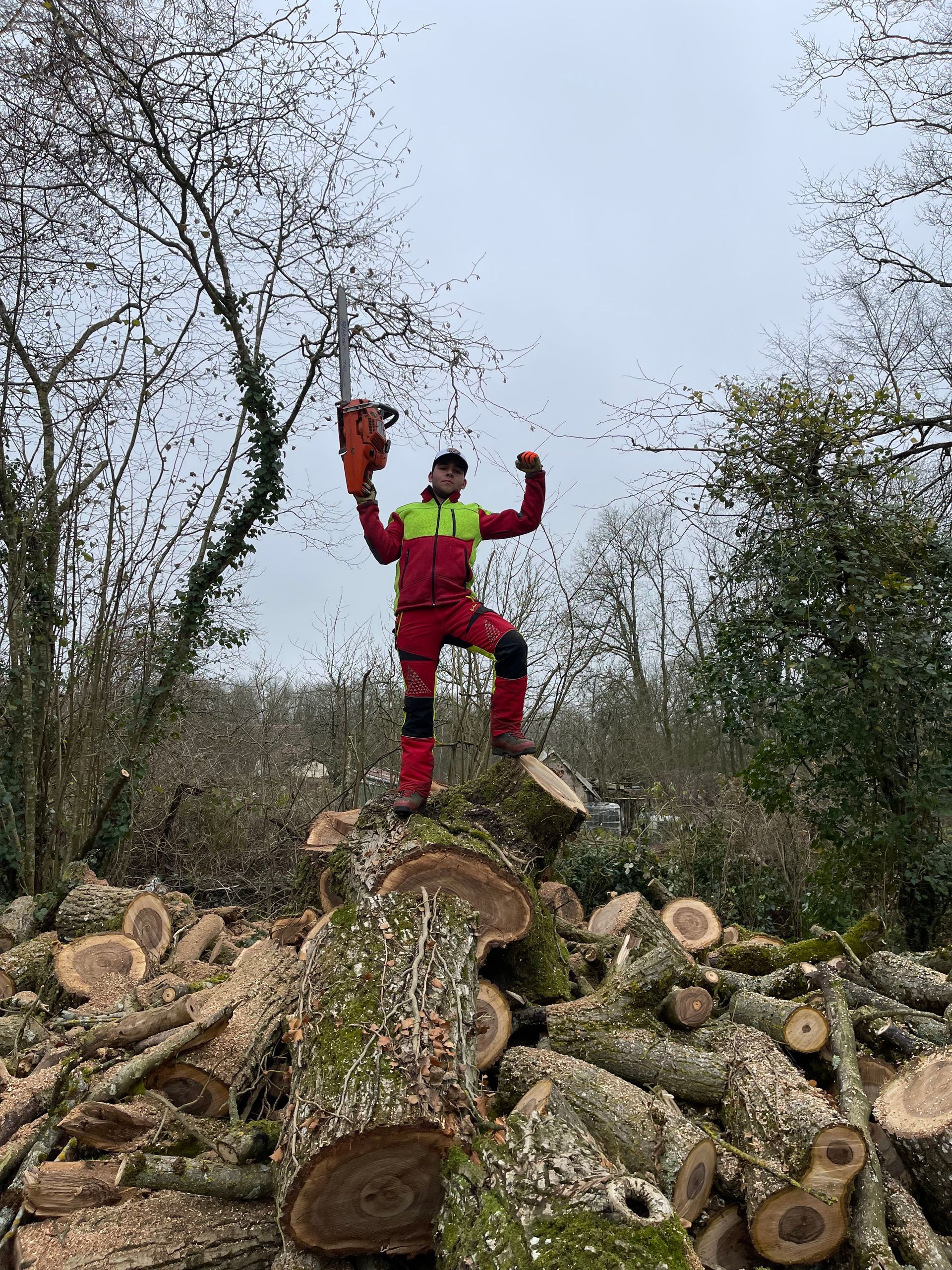 Un homme en vêtements de travail rouges et jaunes se tient debout sur un tas de bûches, brandissant une tronçonneuse, les bras levés dans une pose victorieuse.