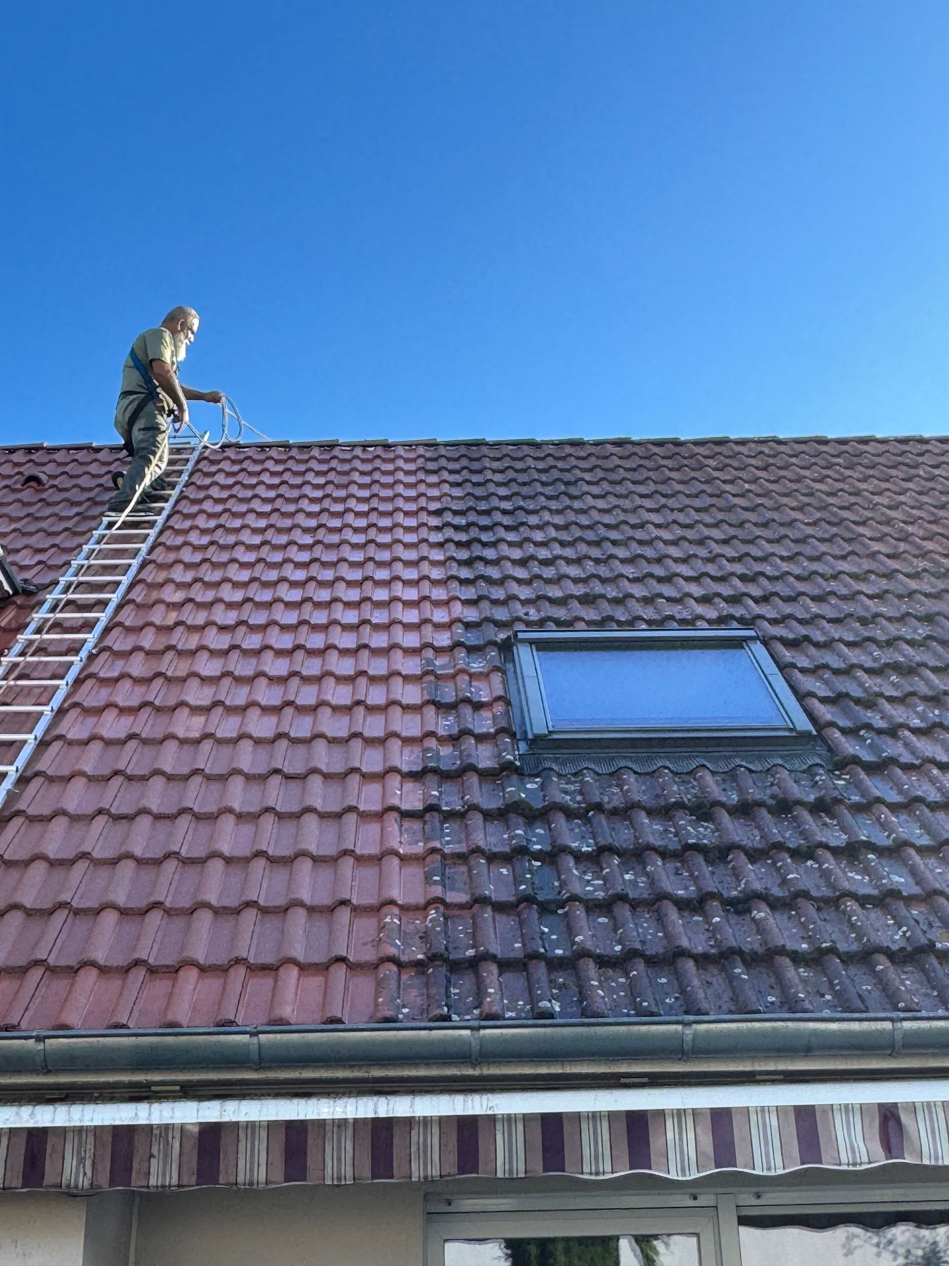 Une personne nettoie à haute pression un toit de tuiles rouges, enlevant la mousse et la saleté ; journée ensoleillée.