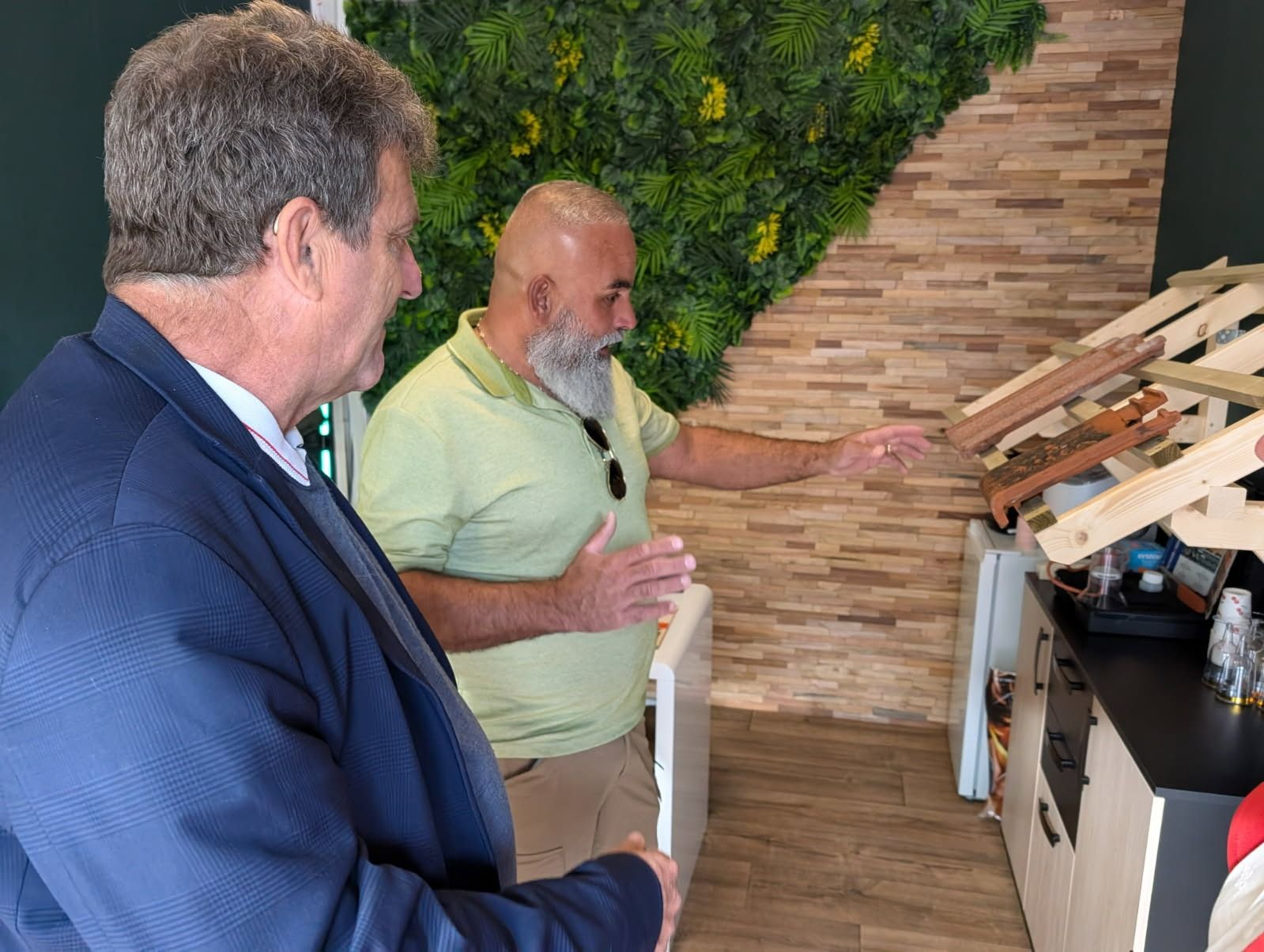 Deux hommes inspectent une petite structure en bois dans une pièce avec un mur vert et des éléments en pierre.