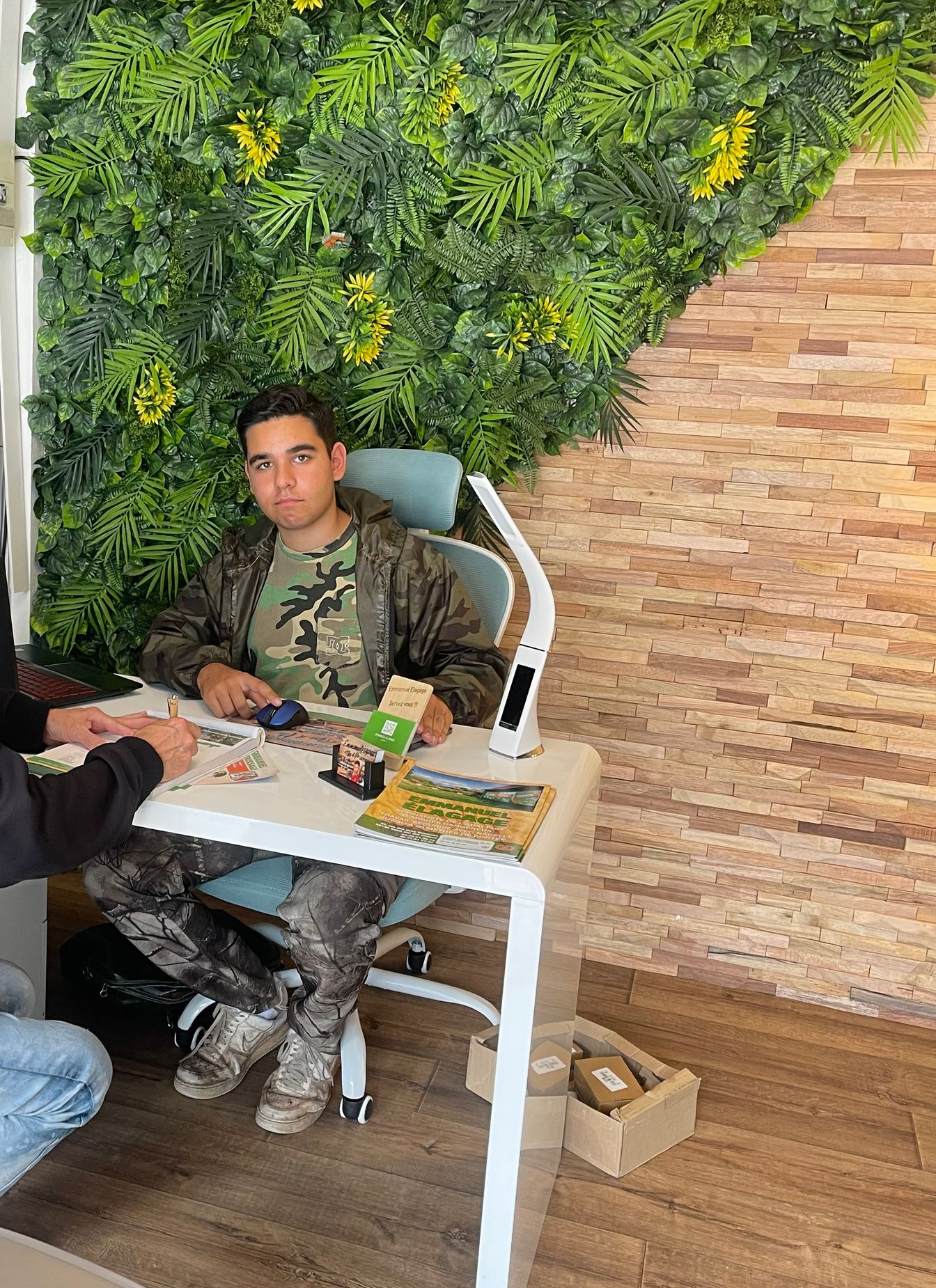 Un homme en tenue de camouflage, assis à un bureau, examine des objets. Un mur vert se trouve derrière lui et un mur de briques sur le côté.