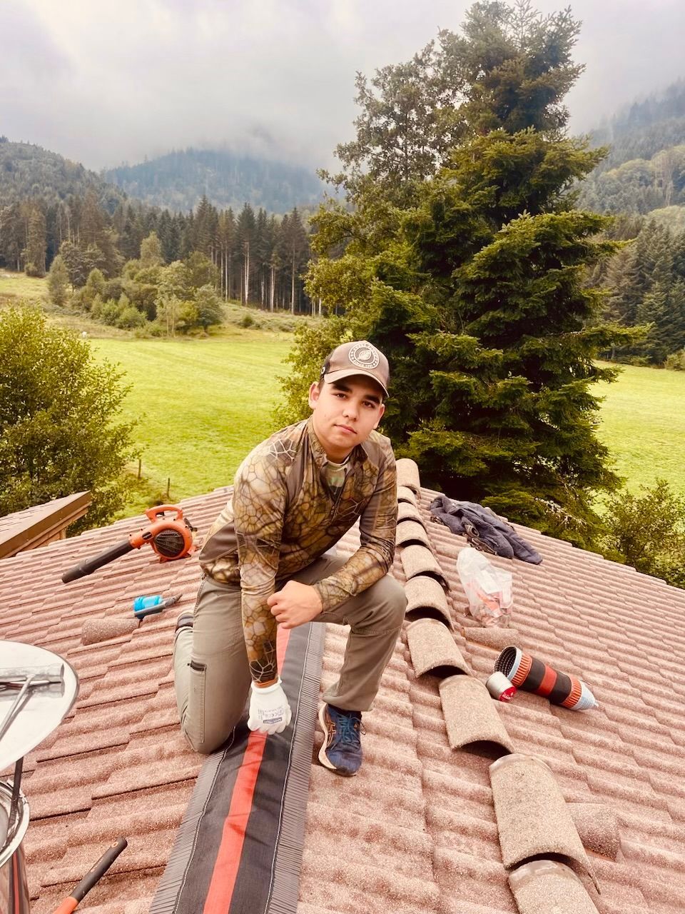 Un homme en tenue de camouflage agenouillé sur un toit de tuiles, avec des montagnes et des arbres en arrière-plan.
