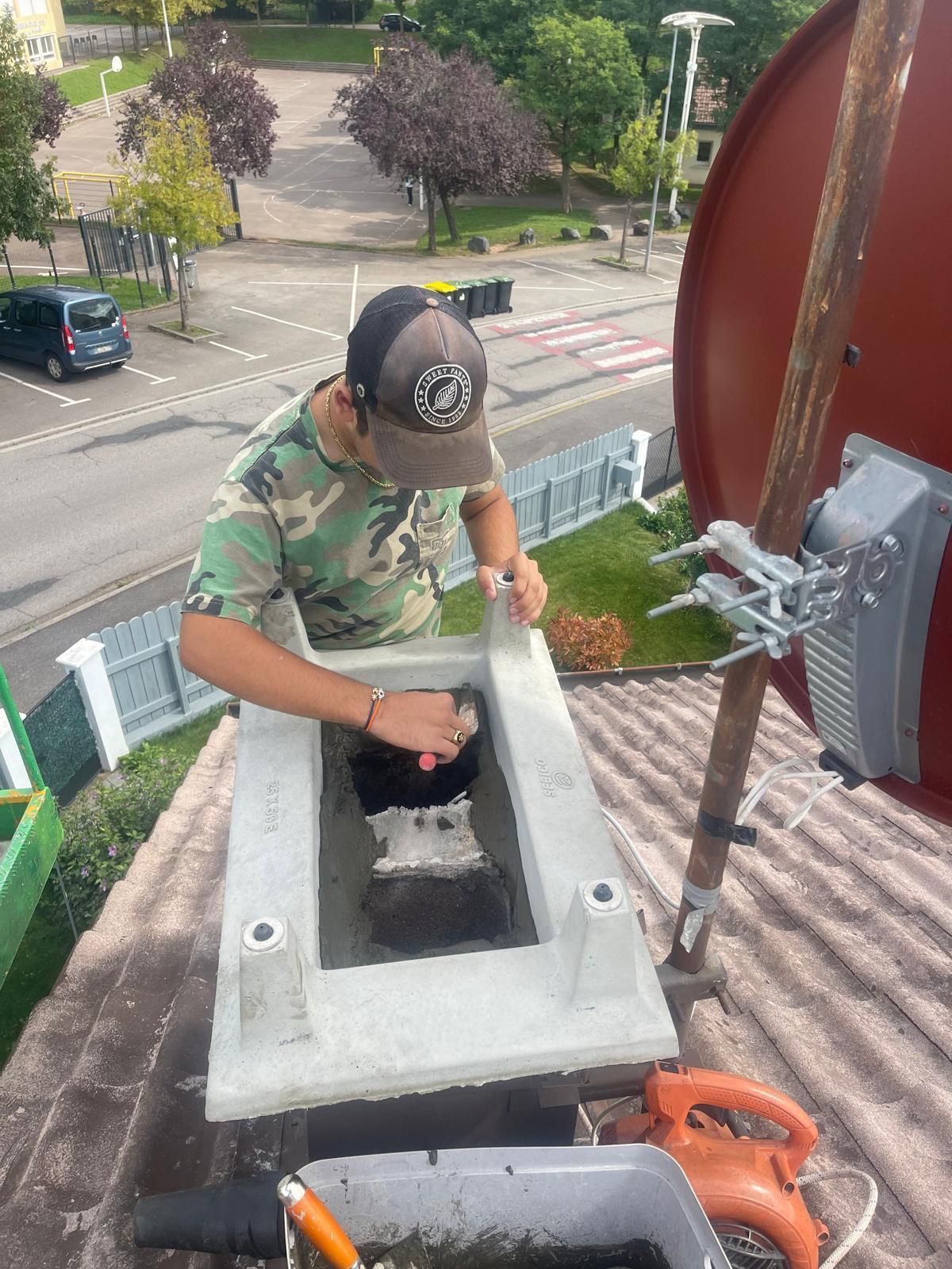 Une personne travaille sur un toit. Elle porte un chapeau et une chemise de camouflage. Antenne parabolique rouge et vue de la rue.