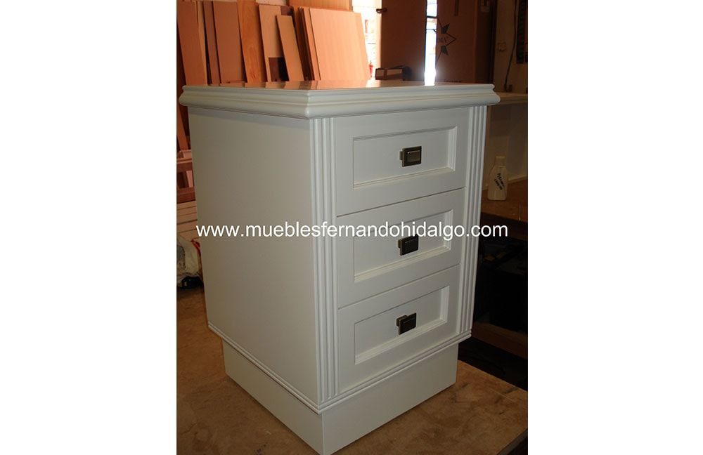 Una mesita de noche blanca con tres cajones y la web www.mueblesfernandohidalgo.com