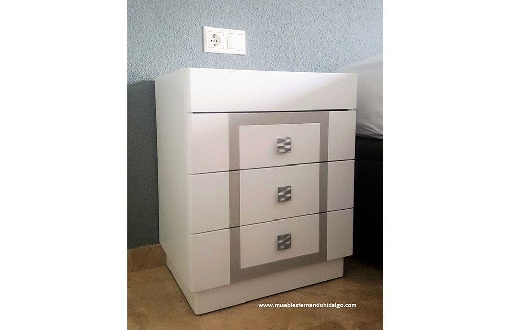 Una mesita de noche blanca con tres cajones está al lado de una cama.