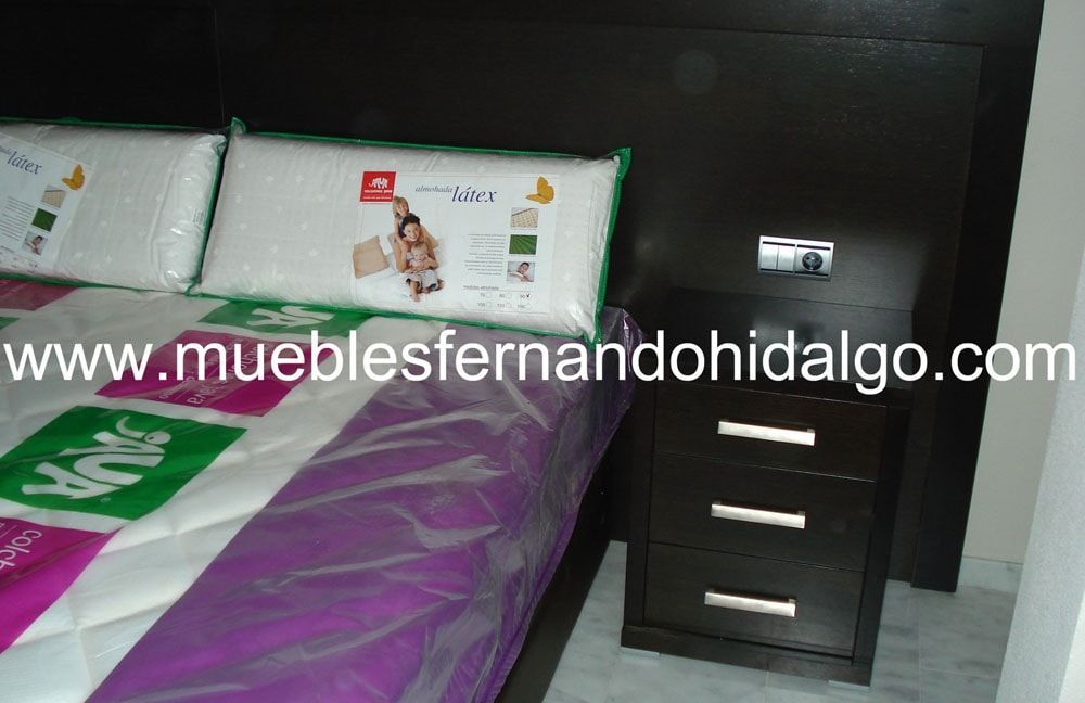 Una cama con manta morada y verde y una mesita de noche con la web www.mueblesfernandohidalgo.com