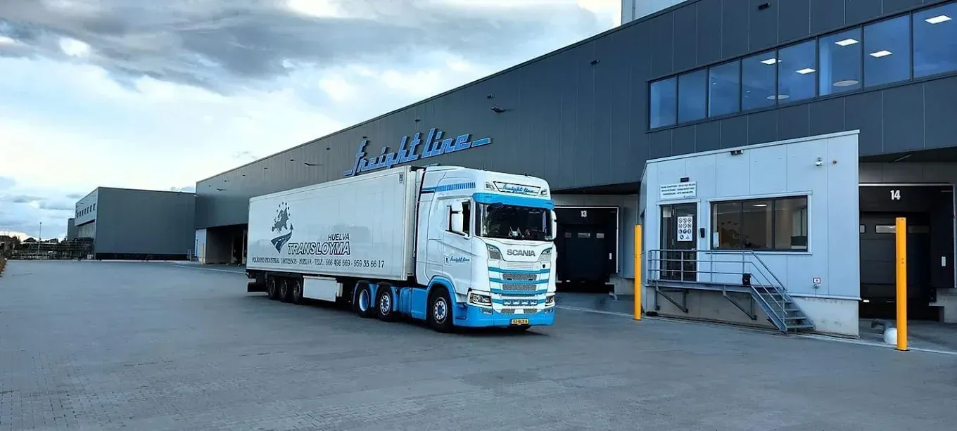 Mujer con jeans y camiseta blanca metiendo la mano en el congelador de un supermercado, sosteniendo una canasta de naranjas.