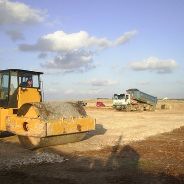 Un rodillo amarillo está sentado en un campo de tierra al lado de un camión volquete.