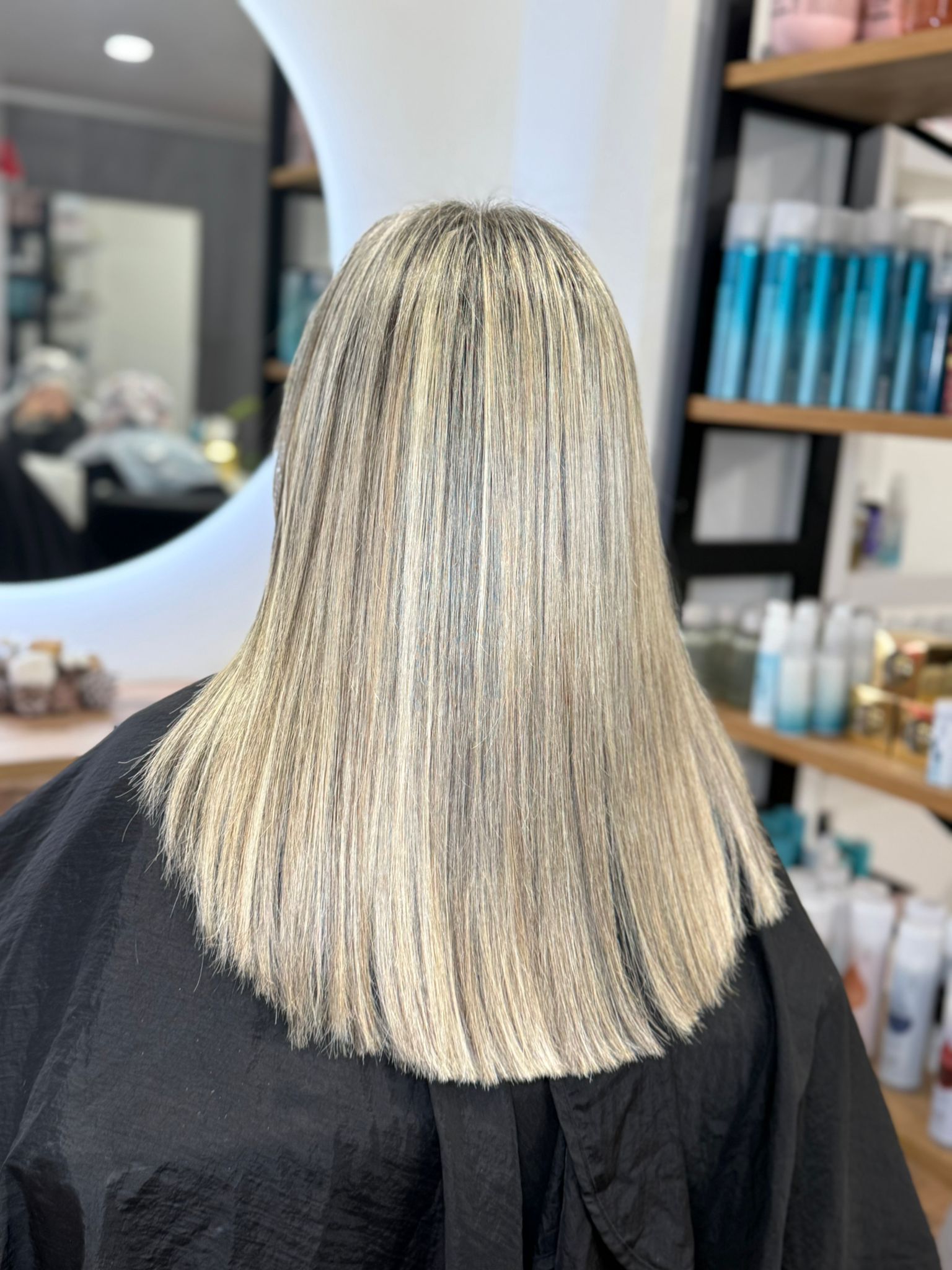 Persona con cabello rubio, liso y en capas. En un salón de belleza, con una capa negra.