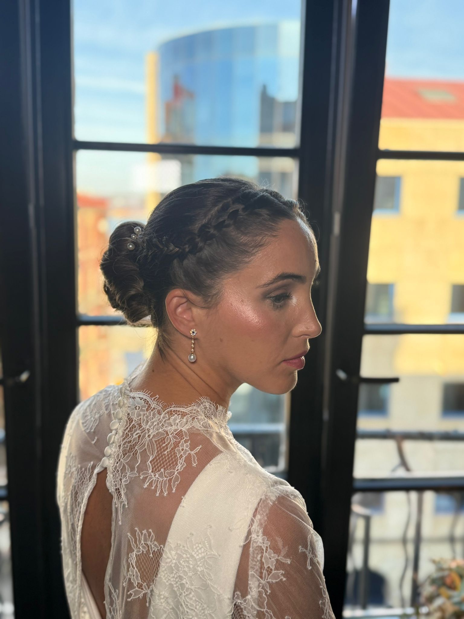 Mujer con moño trenzado y vestido elegante, mirando desde una ventana los edificios de la ciudad.