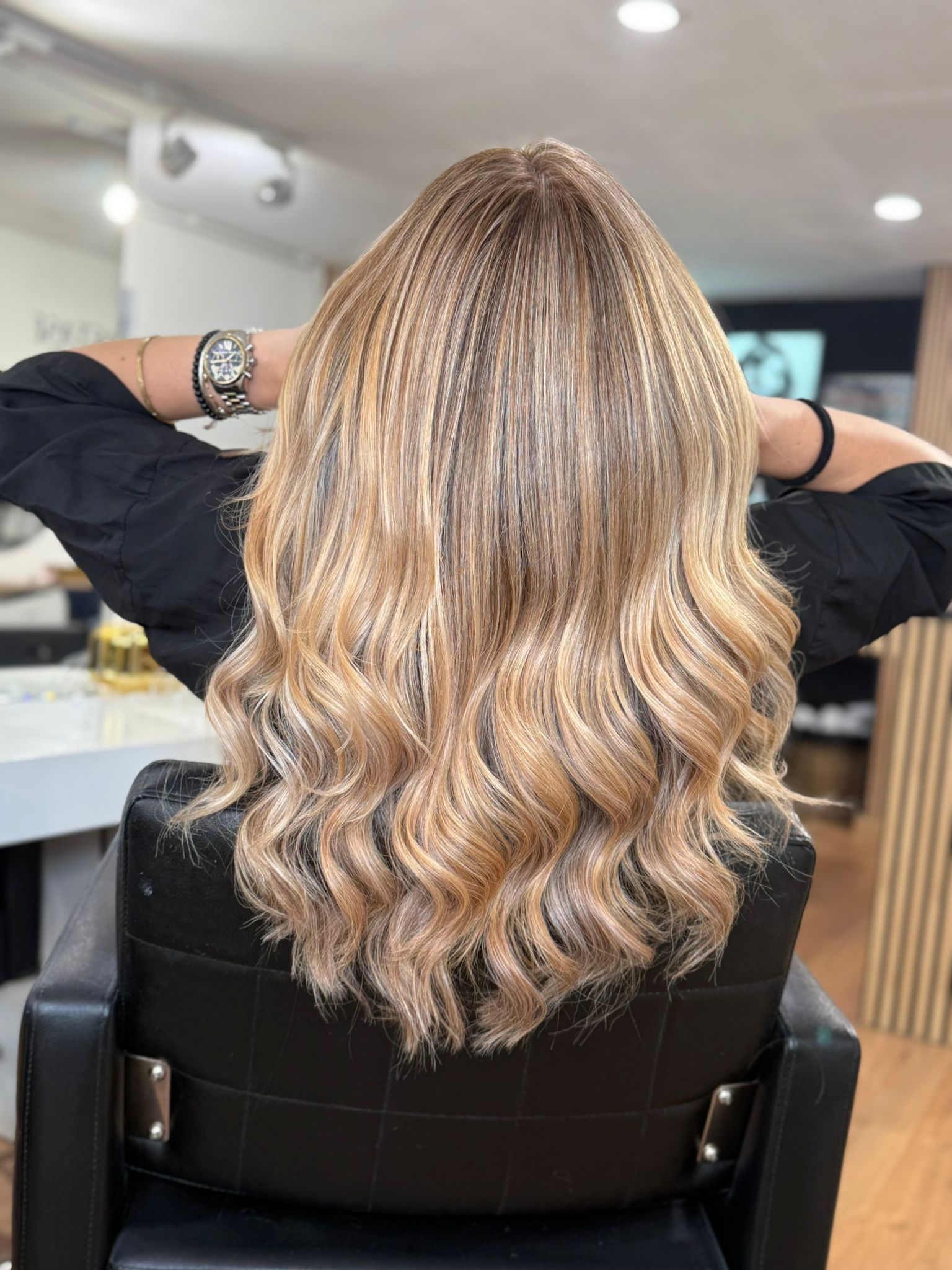 Mujer con cabello rubio ondulado, sentada en una silla de salón con las manos detrás de la cabeza.