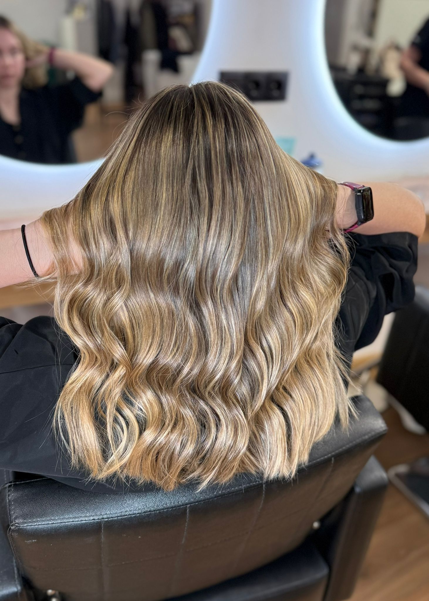 Mujer con cabello largo, ondulado y con reflejos en una silla de salón, con los brazos levantados para tocar el cabello.