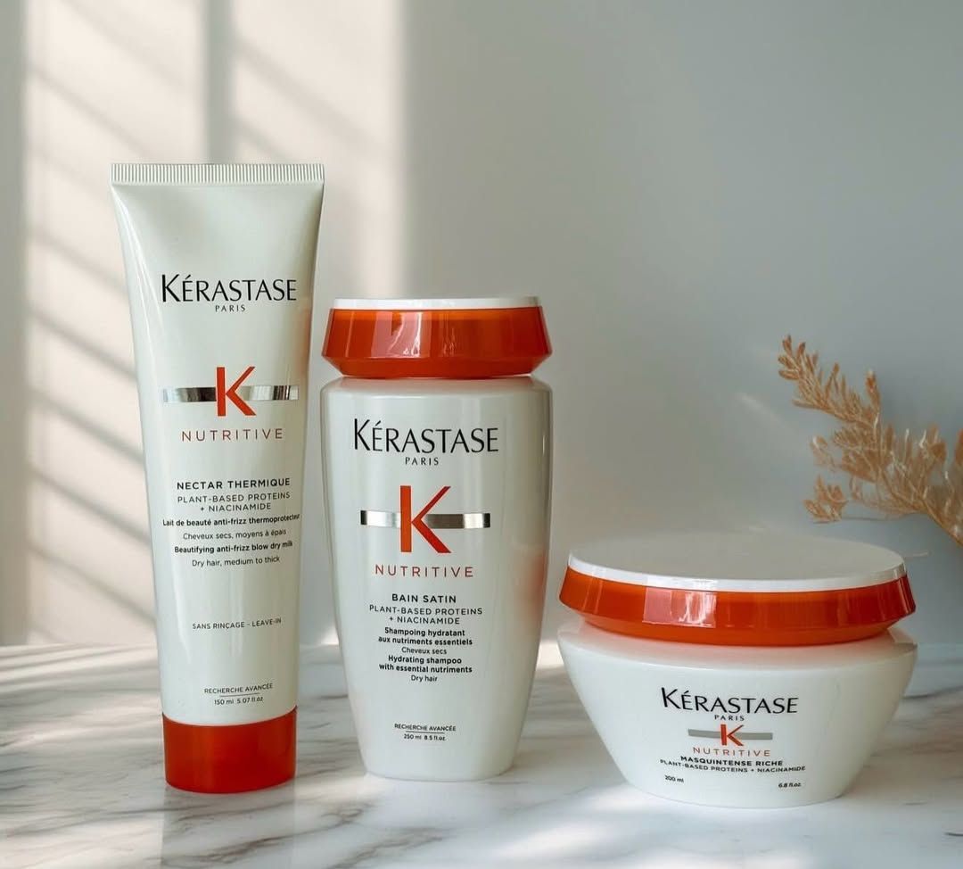 Tres productos para el cuidado del cabello Kerastase, frascos blancos con detalles naranjas, sobre una superficie de mármol.