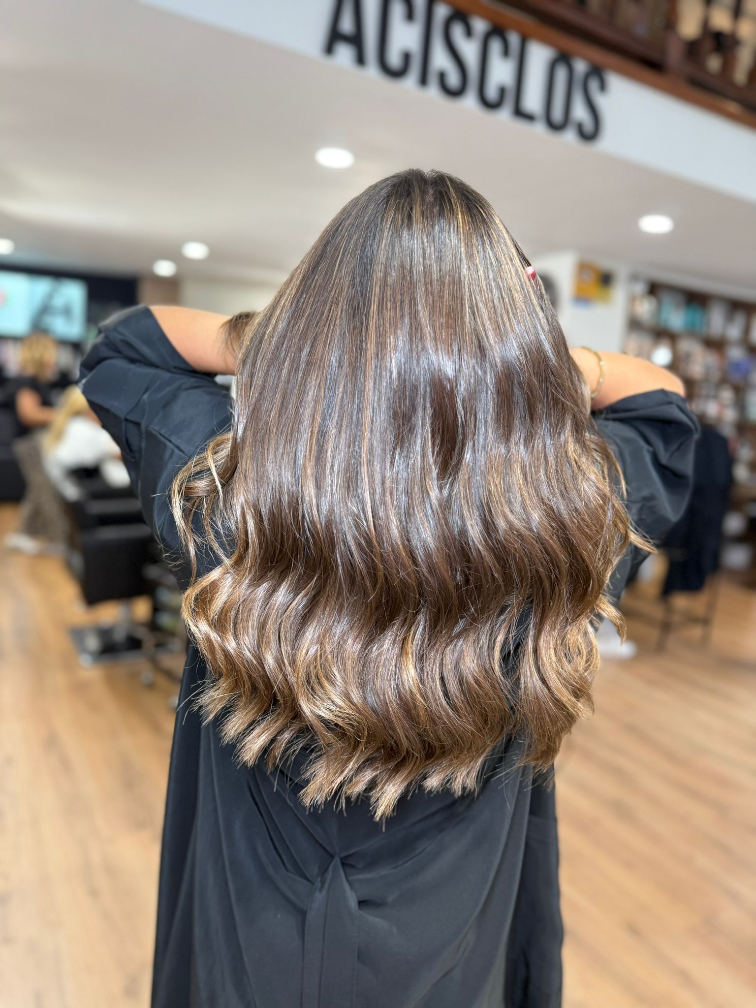 Mujer con cabello castaño largo y ondulado, peinado en un salón, con los brazos levantados.