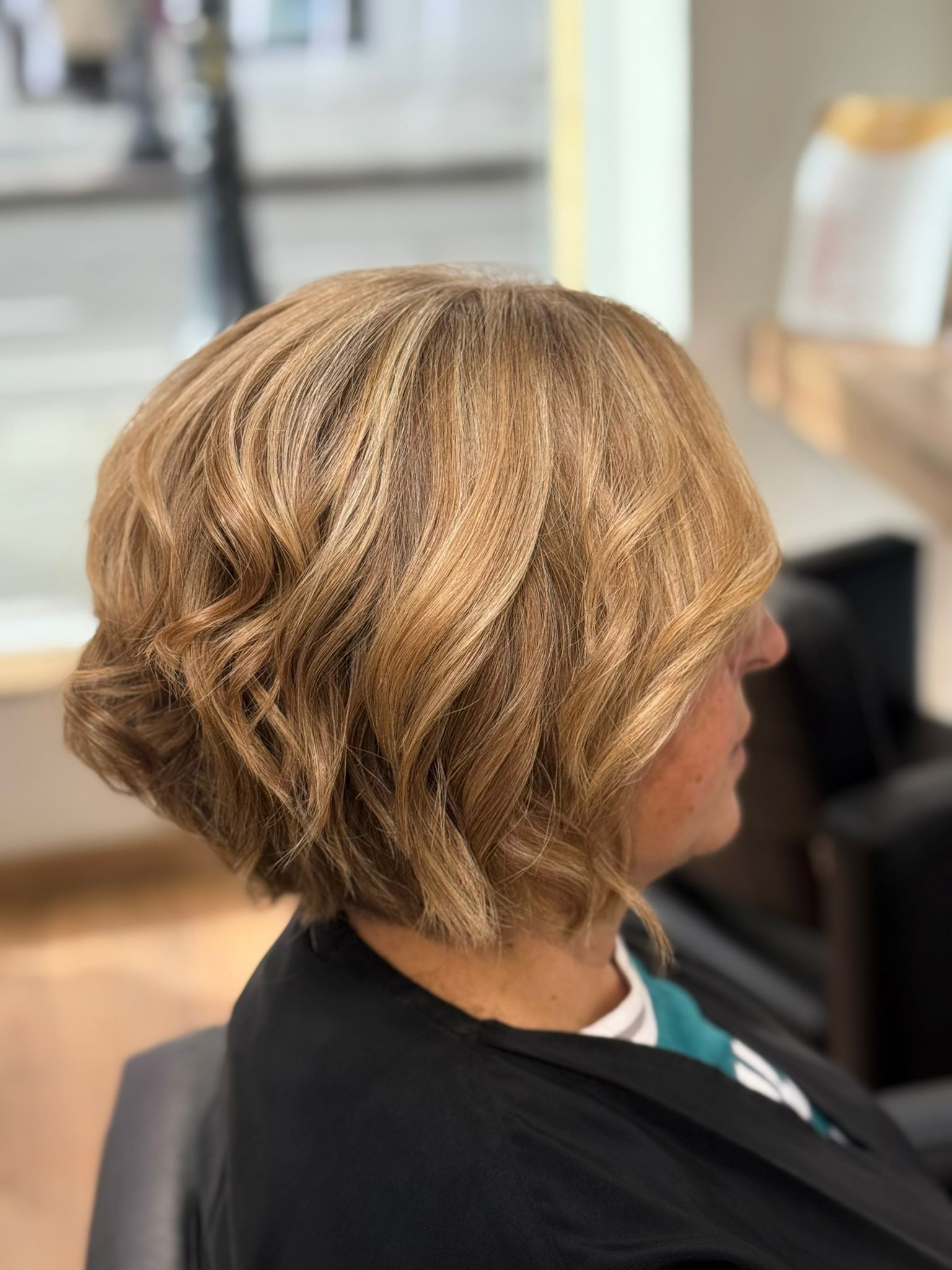 Mujer con cabello rubio ondulado y en capas, vista de perfil. En un salón de belleza.