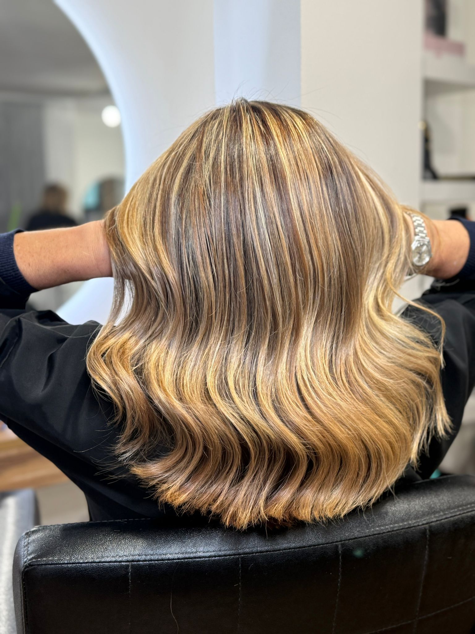 Una mujer con cabello ondulado y resaltado se sienta en una silla de salón, con los brazos detrás de la cabeza.
