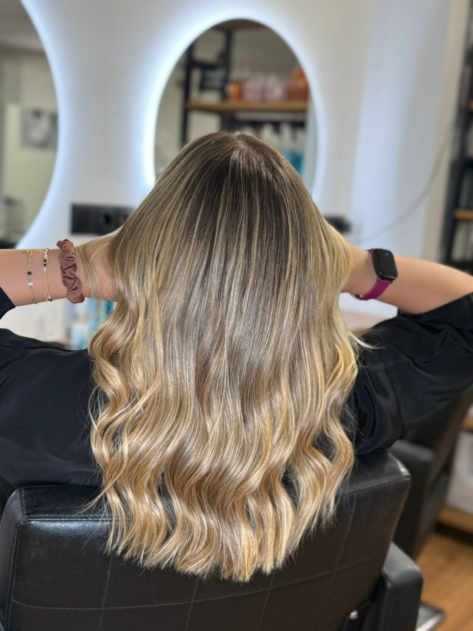 Mujer con cabello rubio ondulado en un salón, raíces oscuras, manos en la parte posterior de la cabeza, sentada en una silla.