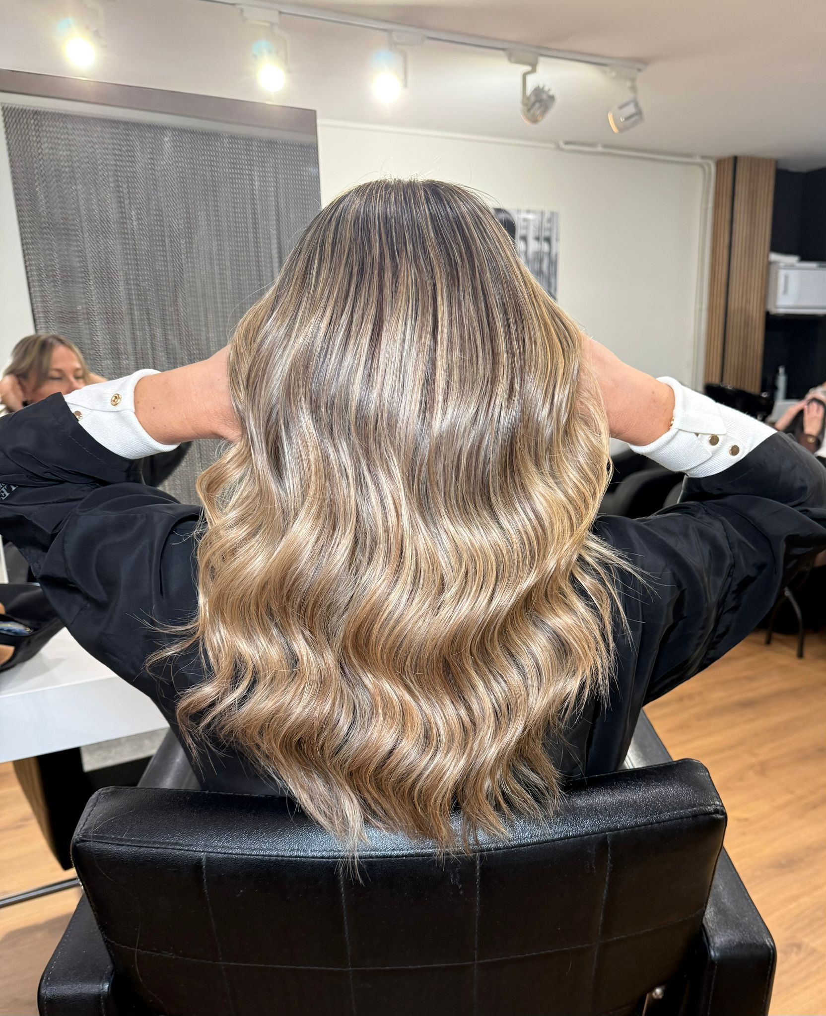 Mujer con cabello rubio ondulado en un salón, sosteniendo las manos detrás de la cabeza, sentada en una silla negra.