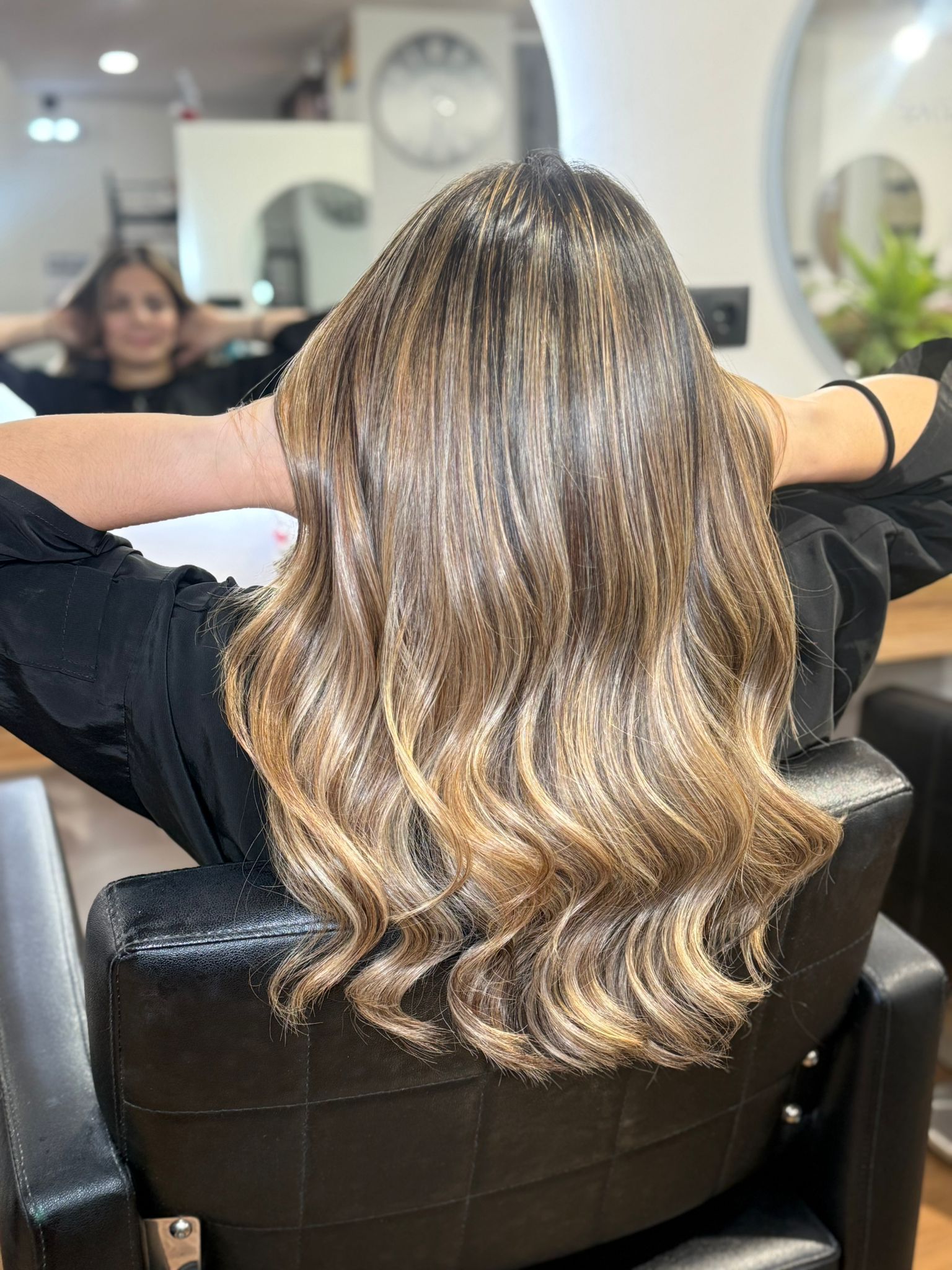 Mujer con cabello largo, ondulado y castaño con reflejos rubios sentada en una silla de salón.