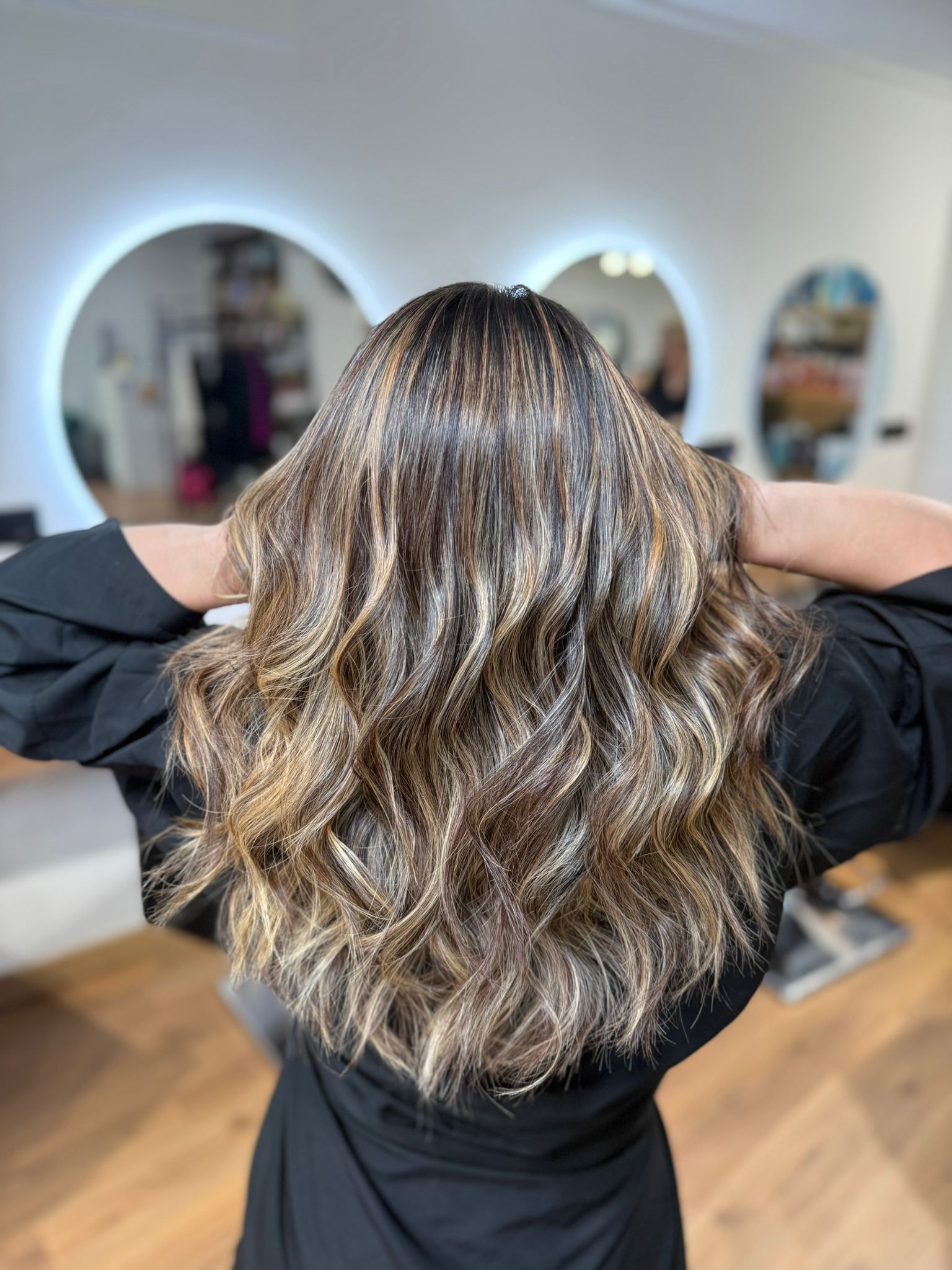 Mujer con cabello castaño ondulado y mechas en un salón de belleza, con las manos en alto. Espejos reflectantes al fondo.