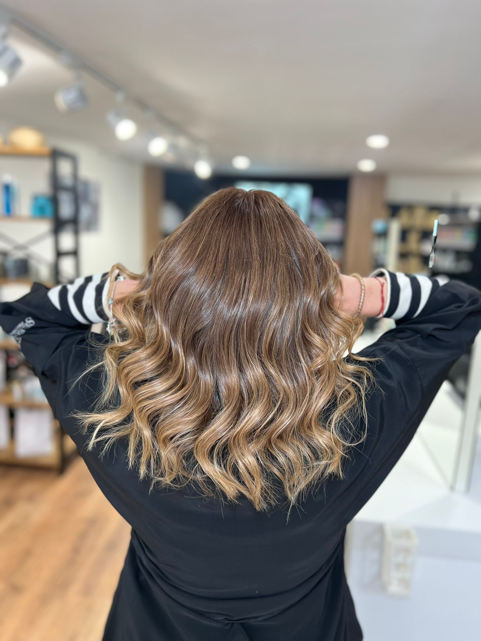Mujer con cabello ondulado y con mechas en un salón, con las manos detrás de la cabeza.