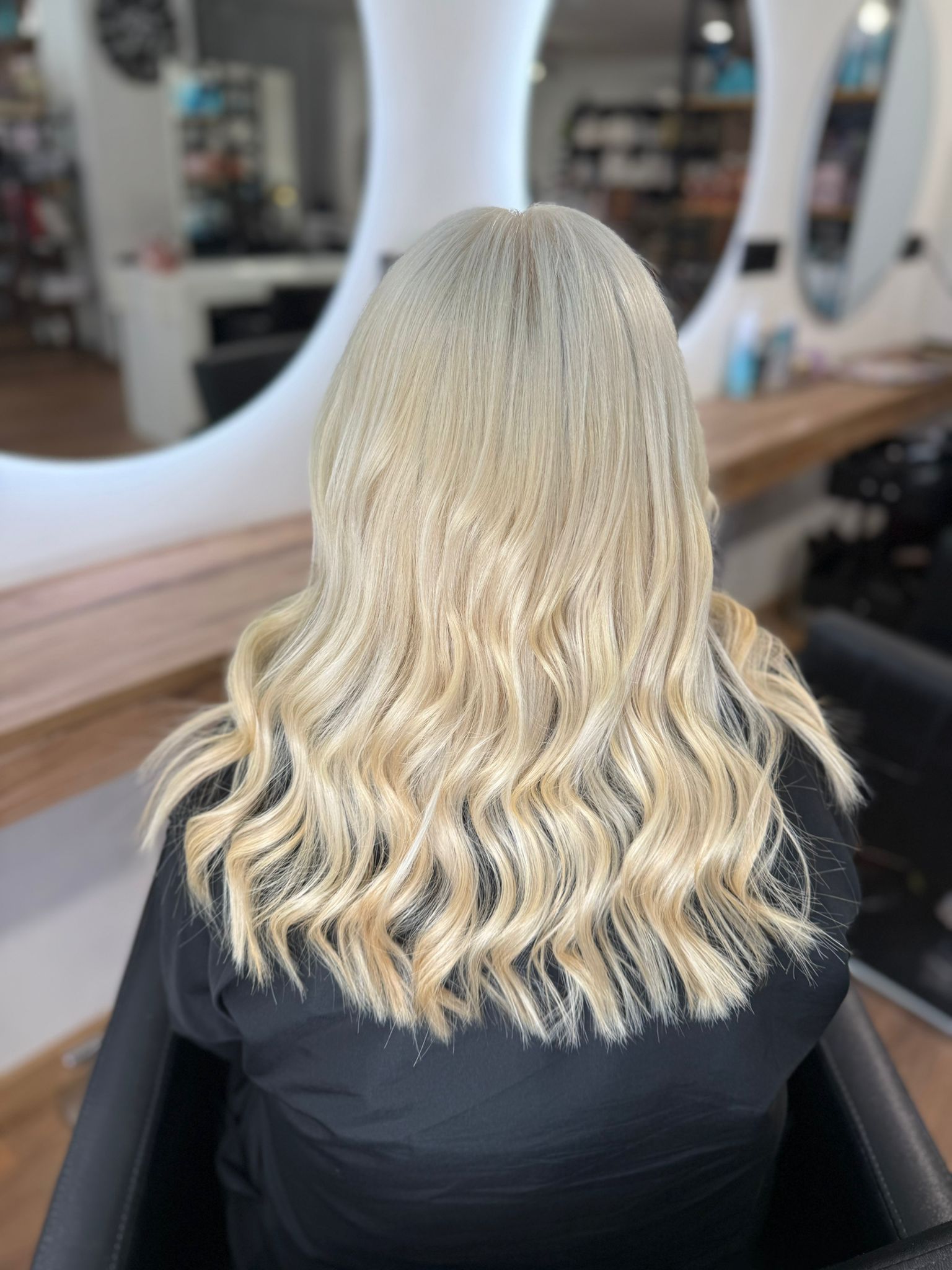 Mujer con cabello largo, ondulado y rubio peinado en un salón, de espaldas a la cámara.