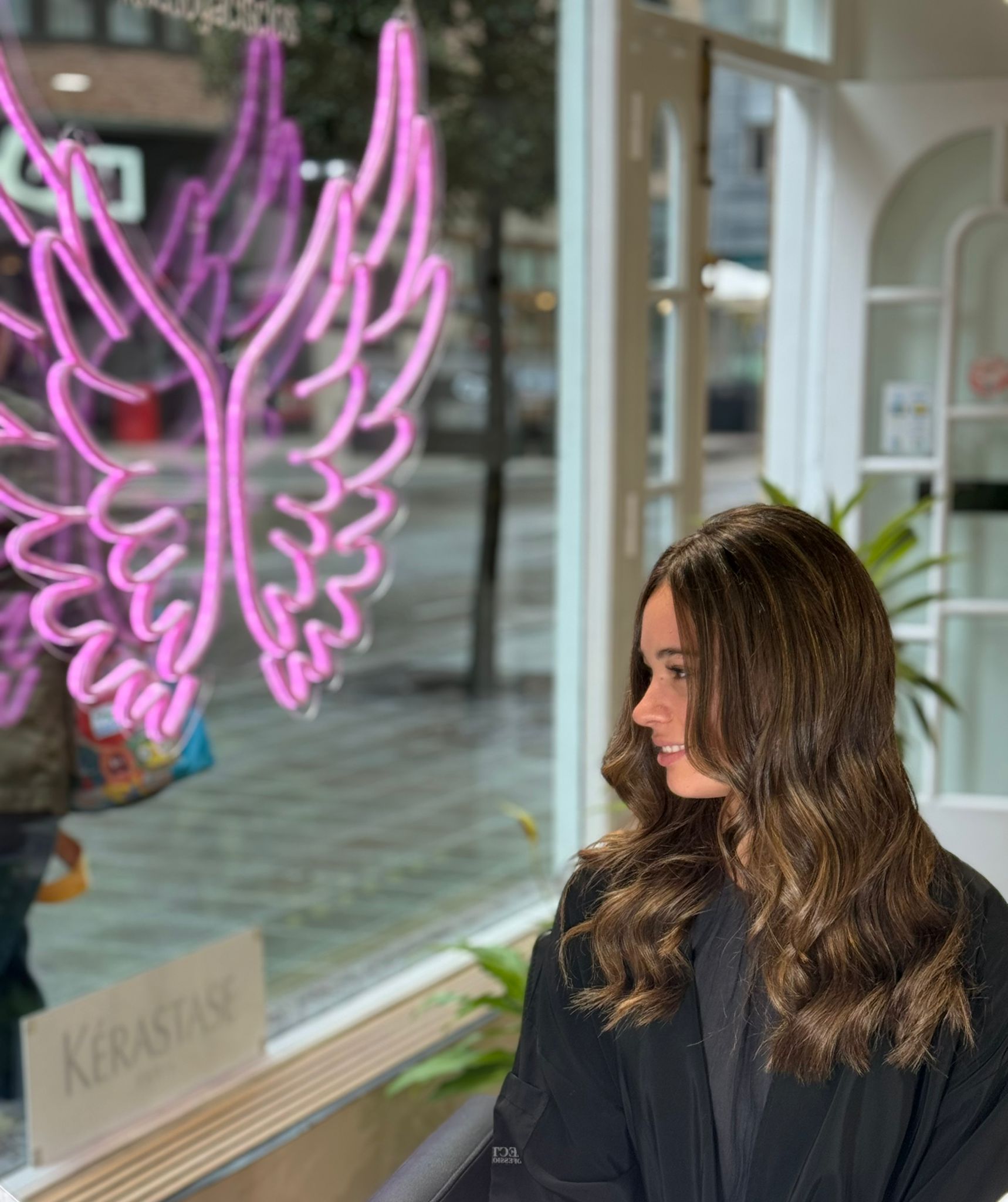 Mujer con cabello castaño ondulado mirando hacia un letrero de ala de ángel de neón rosa en una peluquería.