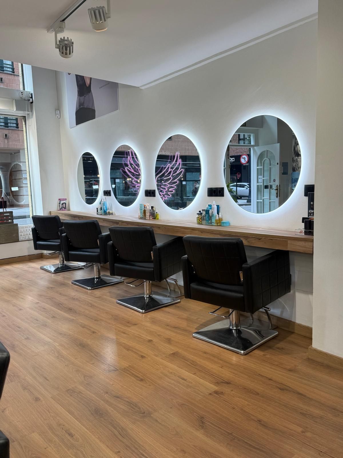 Interior de peluquería con cuatro espejos redondos con iluminación LED sobre sillas negras y pisos de madera.