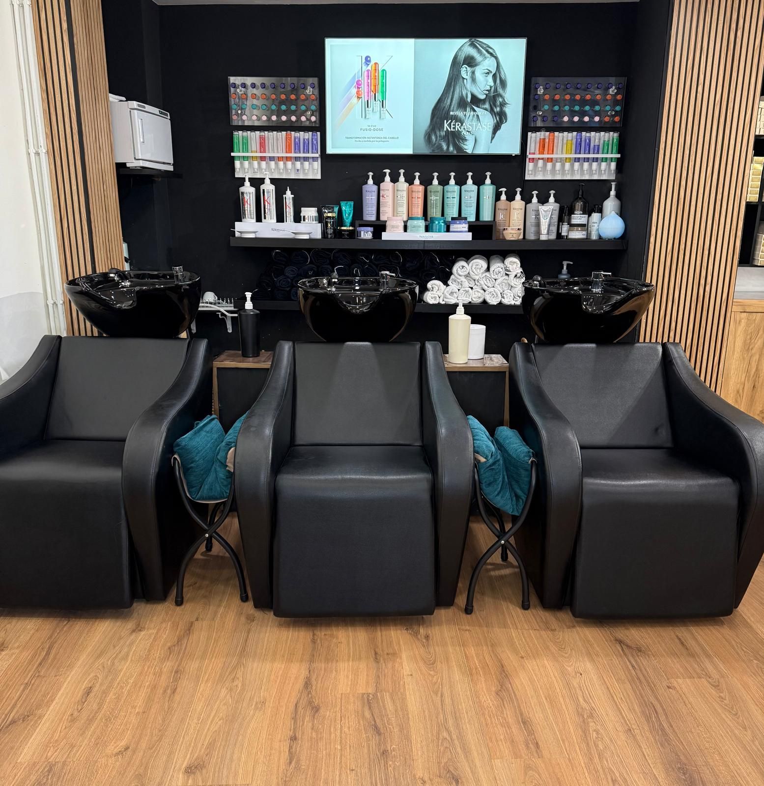 Interior de peluquería con tres sillas negras, lavabos para champú y exhibición de productos.