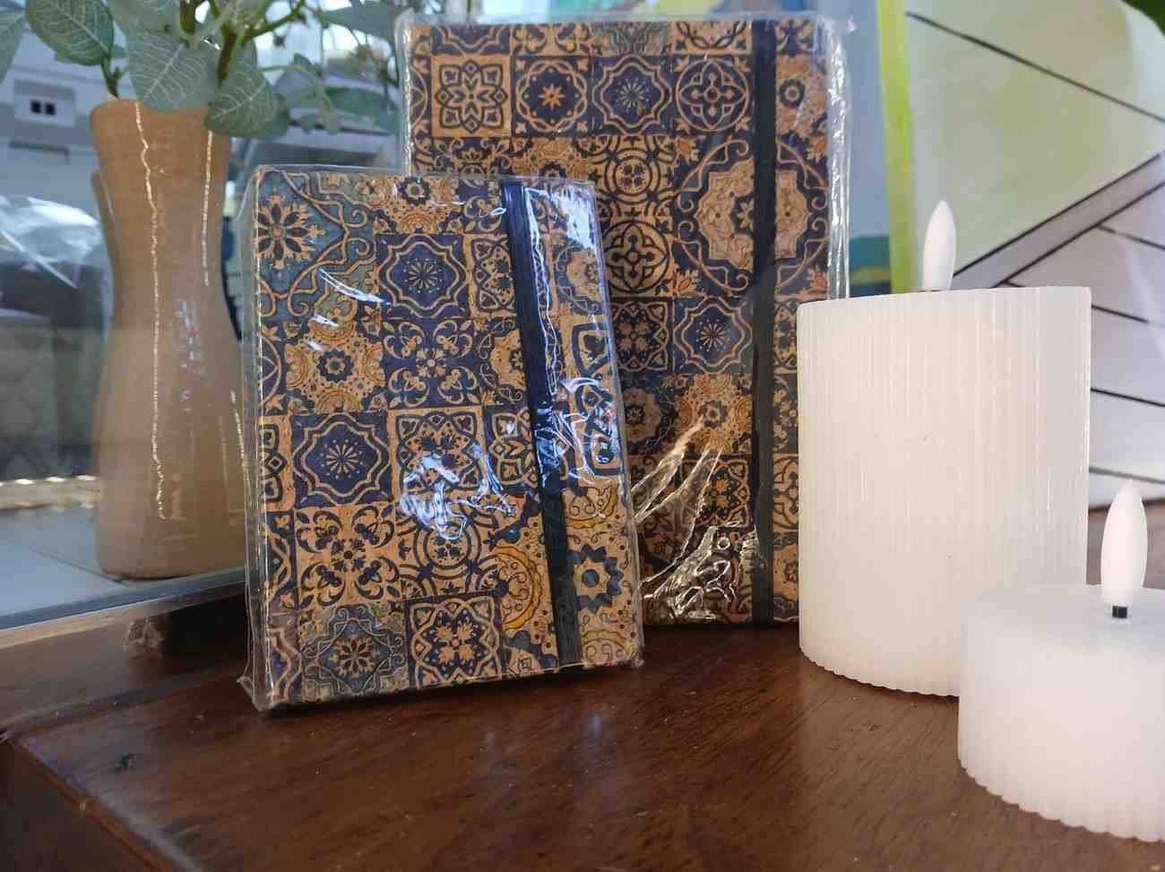 Dos cuadernos estampados con bandas elásticas negras, al lado de dos velas blancas, sobre una superficie de madera.