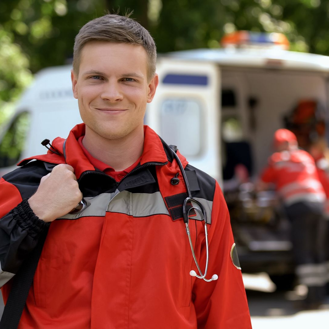 Un ambulancier, souriant devant l'ambulance, porte une veste rouge et son stéthoscope autour du cou.