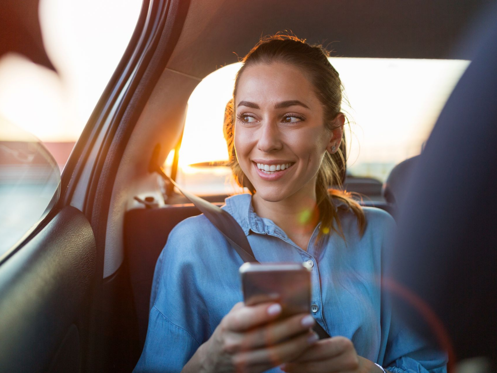Femme souriante, regardant par la fenêtre d'une voiture, tenant son téléphone, coucher de soleil en arrière-plan.