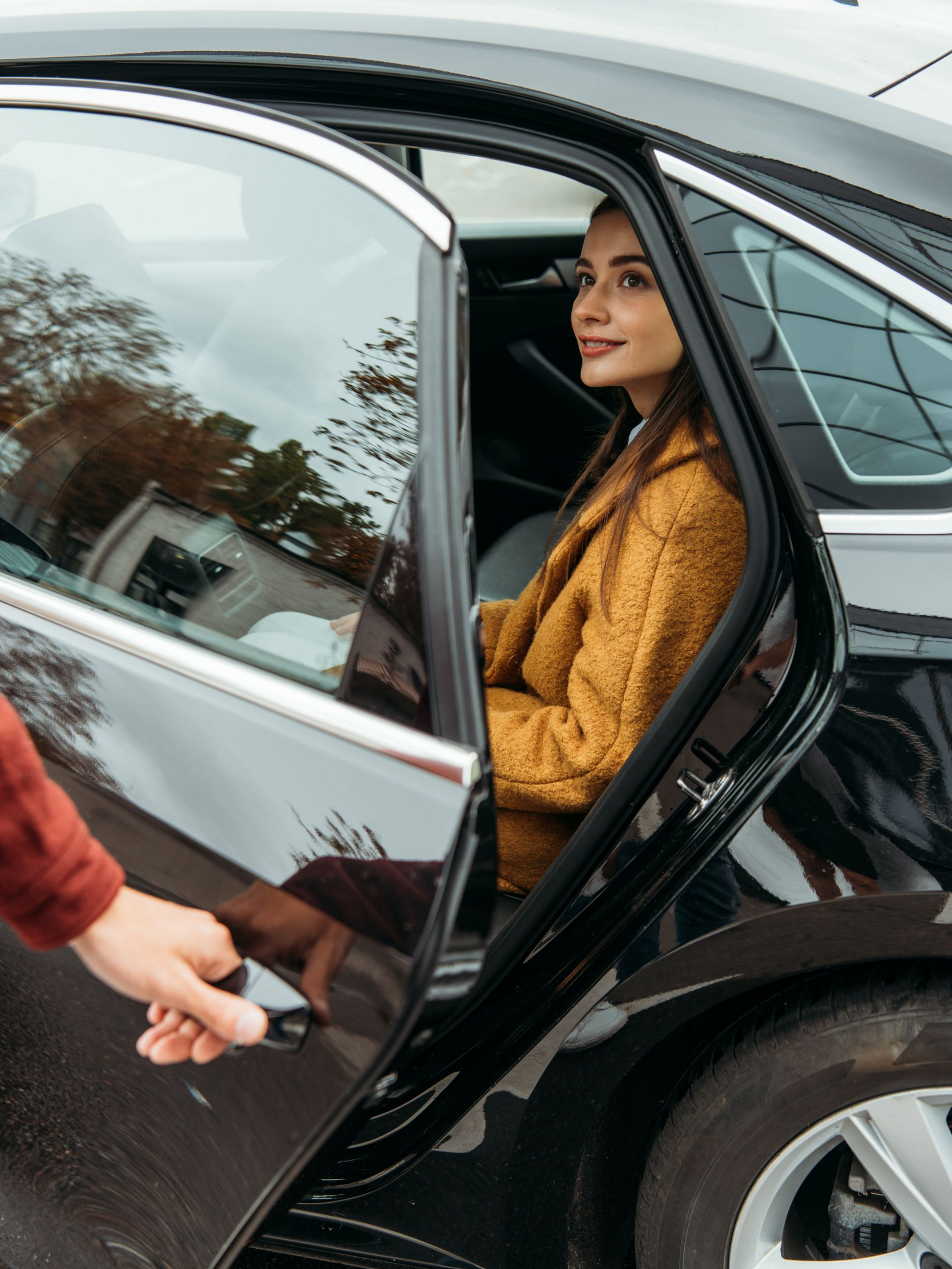 Une femme est assise dans une voiture noire, la portière ouverte, quelqu'un lui ouvre la portière.